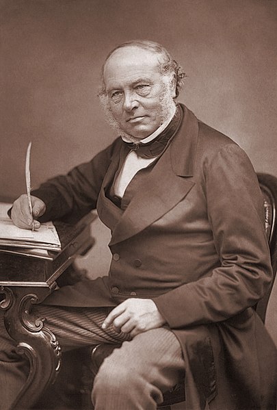 photograph of Sir Rowland Hill, wearing a dark jacket and cravat. He is balding, but has muttonchop sideburns. He is sitting at a desk holding a quill.