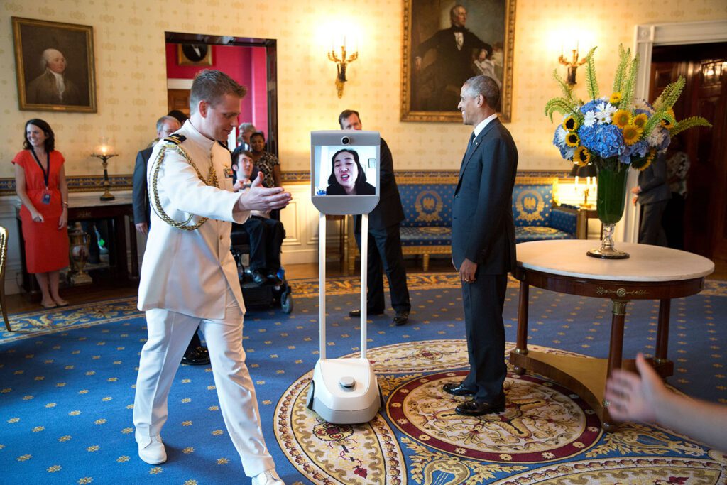 A robot in the middle of a White House room shows a live video of Alice Wong. Wong meets former President Barack Obama, who is standing and smiling to her left. A man in military uniform leads Wong out of the room to her right. There is a line of people standing and in wheelchairs in the background. White House staffers surround Obama and his visitors.