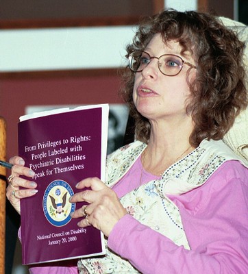 Judi Chamberlin was a middle-aged white woman with brown wavy hair. She wears glasses, a white embroidered vest and pink blouse. She's turned away from the camera, appears to be speaking, and holds the book "From Privileges to Rights: People Labeled with Psychiatric Disabilities Speak for Themselves."