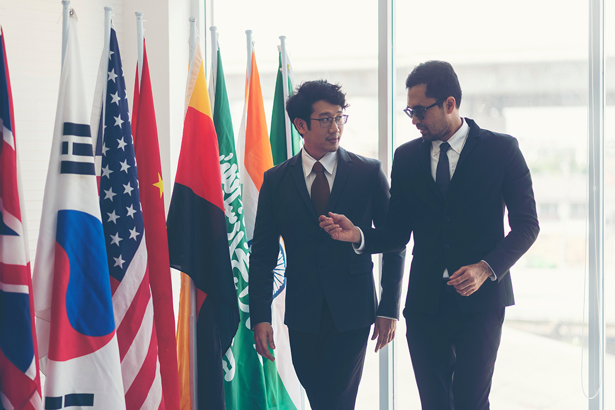 Two men in suits discuss diplomacy near international flags at a global volunteer conference.