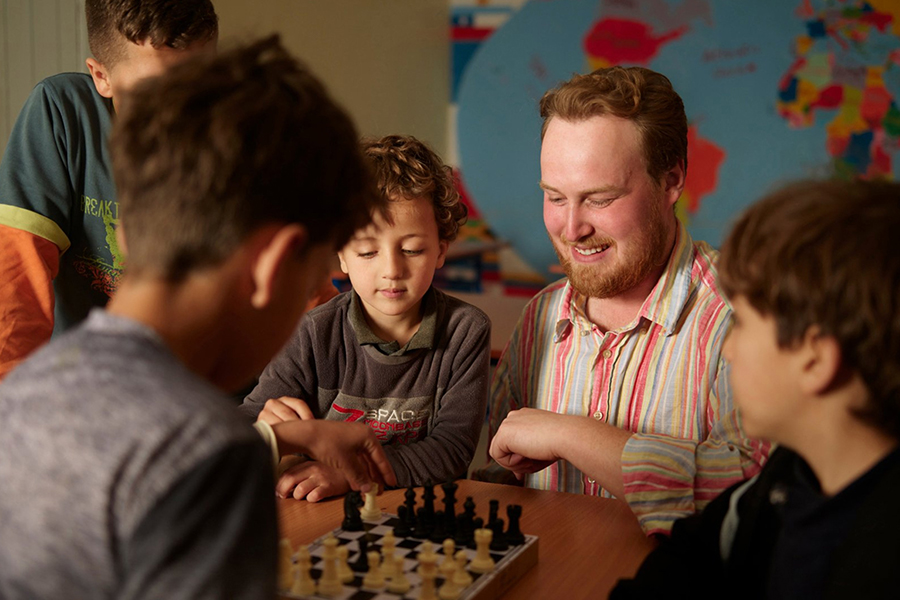 Peace Corps volunteer plays chess with local children while fostering community learning.
