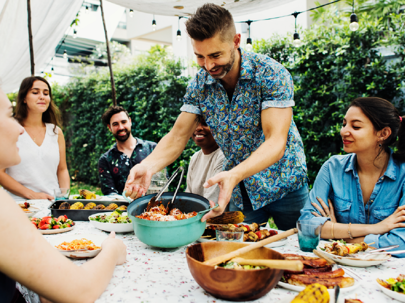 A group of people sitting around a table, as a man is standing and placing a bowl of food on the table